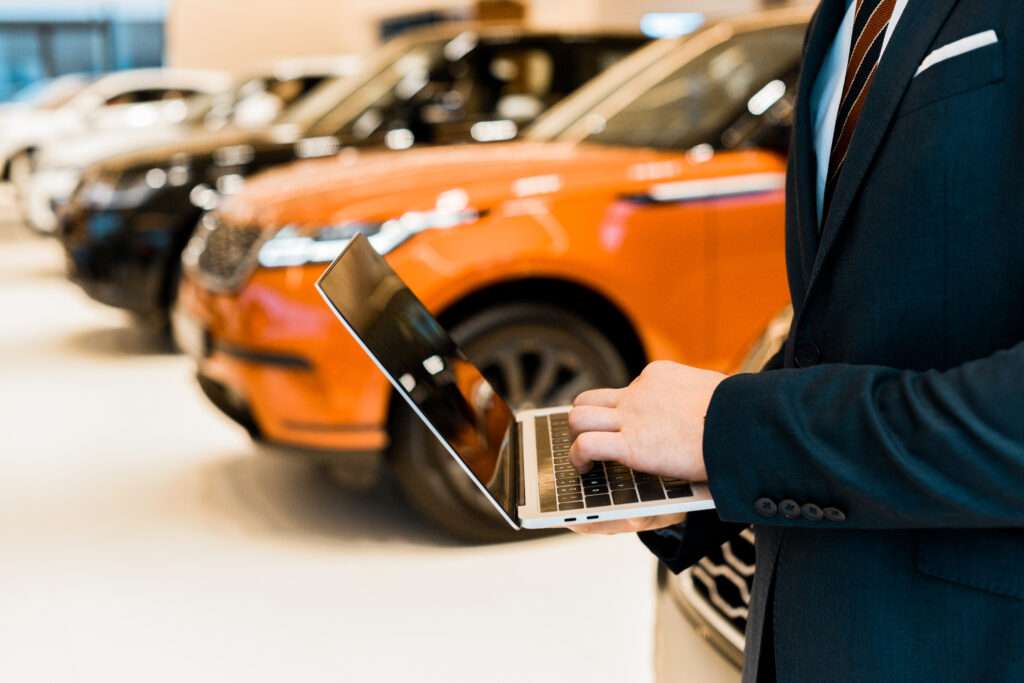 Webdesign Hannover - Eine Person im Anzug benutzt einen Laptop in einem Autohaus und berät möglicherweise Kunden bei der Erstellung von Autohaus-Websites. Im Hintergrund sind mehrere Autos zu sehen, darunter ein orangefarbener SUV. Local SEO Agentur Hannover.