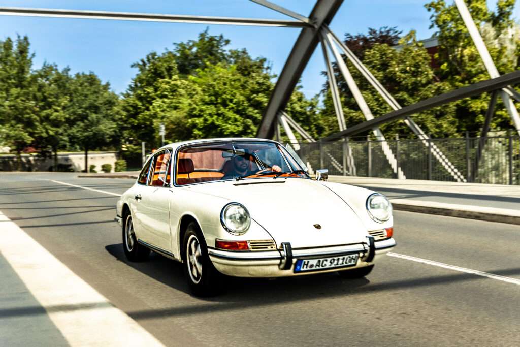 Webdesign Hannover - Ein klassischer weißer Porsche-Sportwagen mit orangefarbener Innenausstattung fährt auf einer Stadtstraße und überquert eine moderne Stahlbrücke – aufgenommen von werbe fotograf hannover – mit Bäumen und Gebäuden im Hintergrund. Local SEO Agentur Hannover.