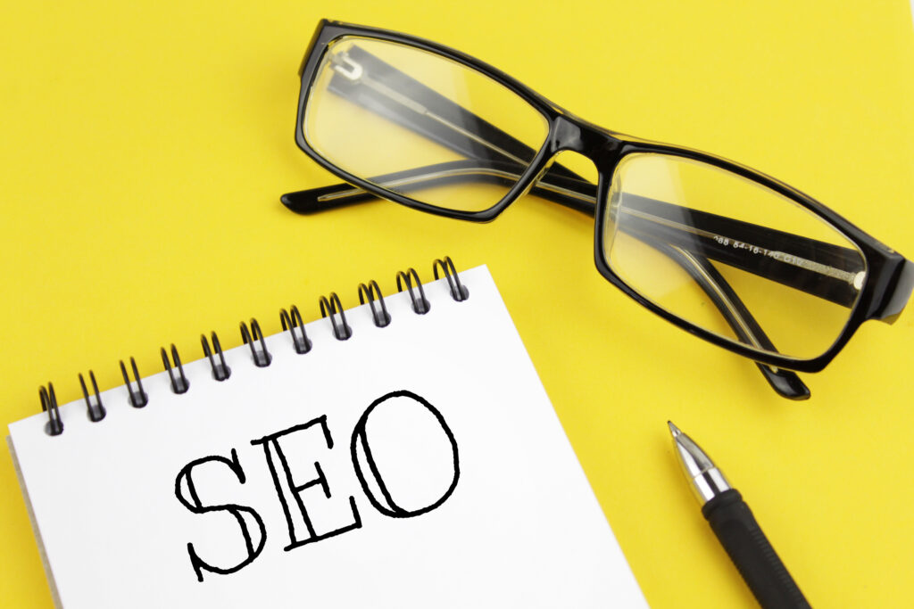 Webdesign Hannover - Eine schwarze Brille, ein schwarzer Stift und ein Notizbuch mit der Aufschrift „SEO“ in großen Buchstaben sind auf einem gelben Hintergrund angeordnet und inspirieren zu Ideen für Google Maps SEO Hannover-Strategien. Local SEO Agentur Hannover.