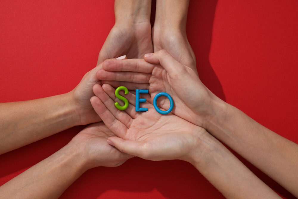 Webdesign Hannover - Mehrere Hände bilden eine Schale und halten Plastikbuchstaben mit der Aufschrift „SEO“ in Blau und Grün auf rotem Hintergrund – perfekt zur Veranschaulichung der SEO-Strategien für Google Maps in Hannover. Local SEO Agentur Hannover.