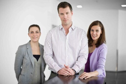 Webdesign Hannover - Drei Personen, zwei Frauen und ein Mann, stehen in legerer Business-Kleidung im Haus. Sie sind der Kamera zugewandt und lächeln leicht, während im Hintergrund neutrale weiße Wände zu sehen sind. Local SEO Agentur Hannover.