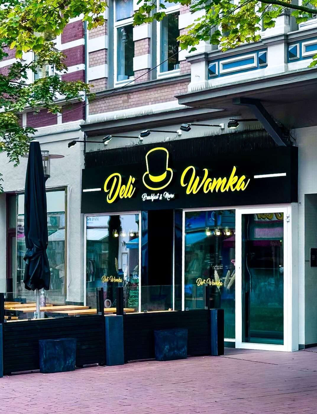 Webdesign Hannover - Ein Schaufenster mit einem schwarzen Schild, auf dem in gelber Schrift "Deli Womka" steht. Ihr einzigartiges Logo mit einem gelben Zylinder ist ein gutes Beispiel für diejenigen, die ein Logo erstellen lassen wollen. Draußen stehen Holzbänke und Tische mit einem geschlossenen schwarzen Regenschirm. Local SEO Agentur Hannover.
