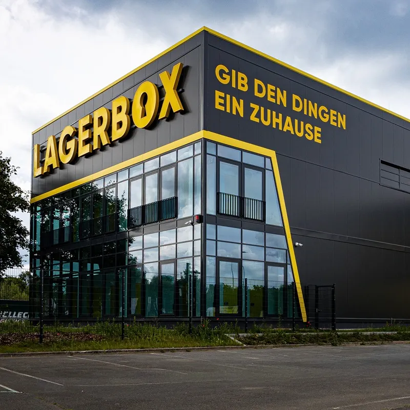 Webdesign Hannover - Ein modernes schwarzes Gebäude mit gelben Zierleisten, das sich perfekt für einen Werbefotografen eignet, trägt die Aufschriften „LAGERBOX“ und „GIB DEN DINGEN EIN ZUHAUSE“ in kräftigen gelben Buchstaben. Das Gebäude verfügt über große Glasfenster und steht neben einem leeren Parkplatz. Local SEO Agentur Hannover.